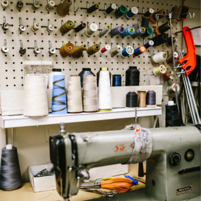 A sewing machine sits on a worktable in front of a wall with spools of thread, scissors, and various sewing tools organized on pegs and shelves.