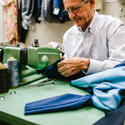 At Teds Clothiers, an experienced tailor uses a sewing machine to perform Pant Hem Alterations, expertly stitching blue fabric with threads and sewing tools on hand.