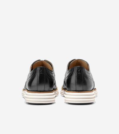 A pair of Cole Haan Grand Remastered Wingtip Oxford black leather shoes with brogued upper and white FlowerFoam midsole by Ted's Clothiers, viewed from the back on a plain white background.