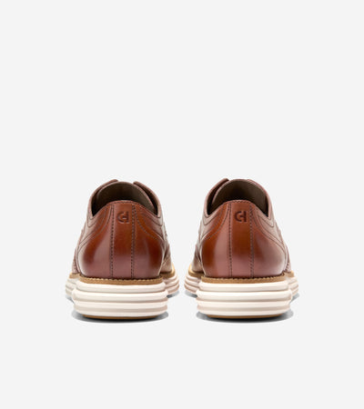 A pair of Cole Haan Grand Remastered Wingtip Oxfords in brown leather with broguing, white soles, and a FlowerFoam midsole from Ted's Clothiers, shown from the back on a plain white background.
