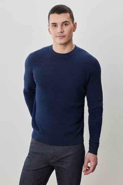 A man faces forward wearing a fitted navy blue Robert Barakett Watertown Sweater by Robert Barakett and dark jeans against a plain light background.