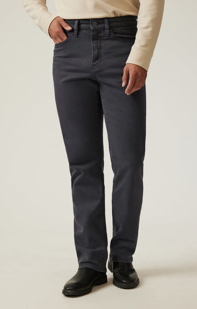 A person wearing a beige long-sleeve shirt, 34 Heritage Ash Twill straight leg trousers by 34 Heritage in dark gray, and black boots stands against a plain light background.
