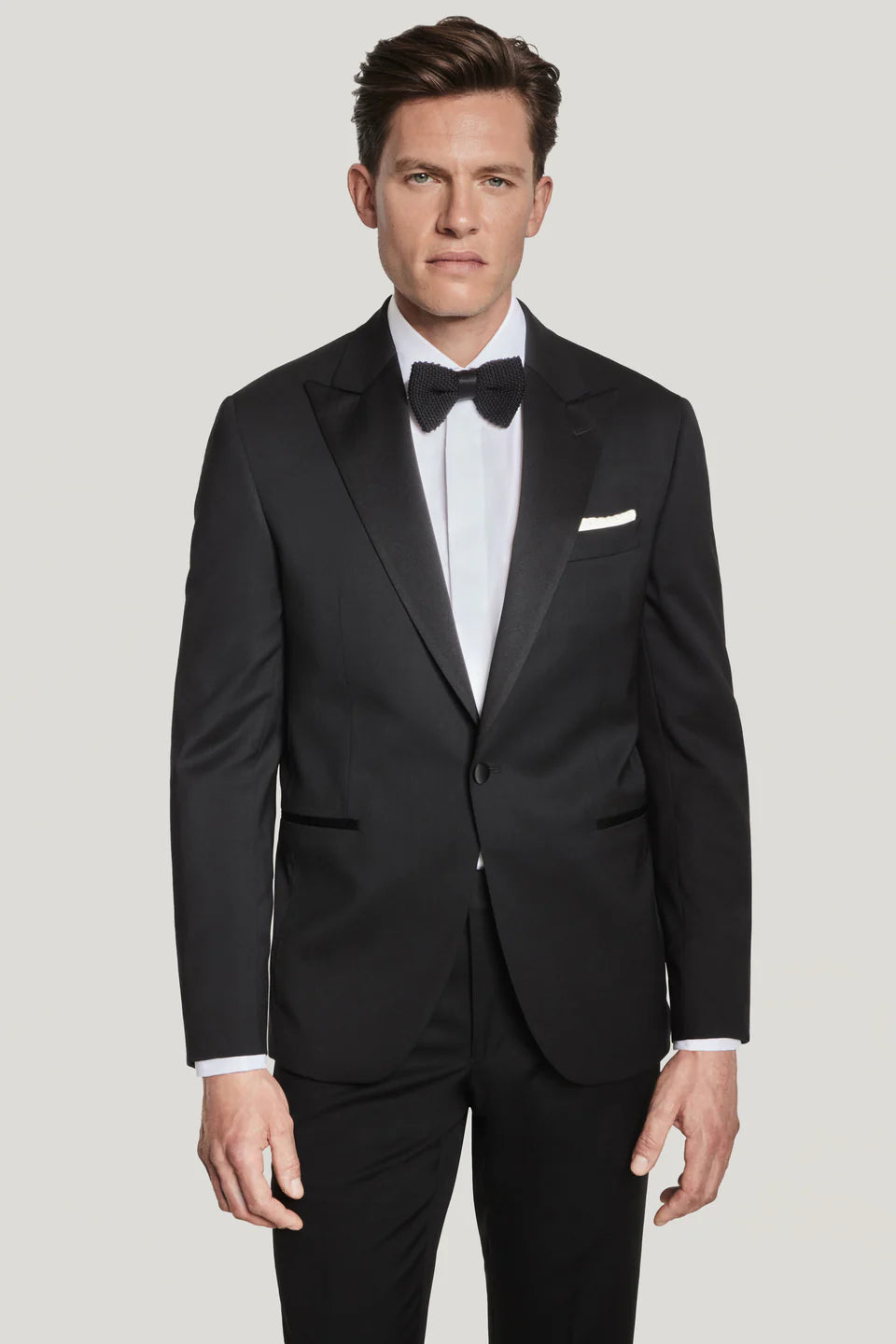 A man wearing a Jack Victor Elwood Solid Tuxedo with a peak lapel, white dress shirt, and black bow tie stands facing forward against a plain light background.