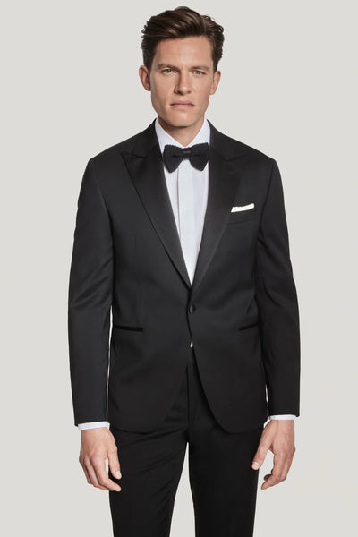 A man wearing a Jack Victor Elwood Solid Tuxedo with a peak lapel, white dress shirt, and black bow tie stands facing forward against a plain light background.