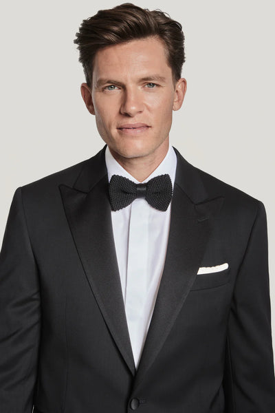 A man wears the Jack Victor Elwood Solid Tuxedo, featuring a peak lapel and crafted from Super 130's wool, paired with a white shirt, black bow tie, and white pocket square, standing against a plain background.