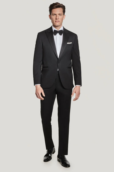 A man stands against a plain light background wearing the Jack Victor Elwood Solid Tuxedo with a peak lapel, crafted from luxurious Super 130's wool, paired with a white dress shirt, black bow tie, pocket square, and black dress shoes.