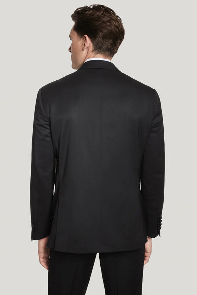 A man in a Jack Victor Elwood Solid Tuxedo with a peak lapel stands facing away from the camera, showcasing the back of his Super 130's wool jacket against a plain light background.