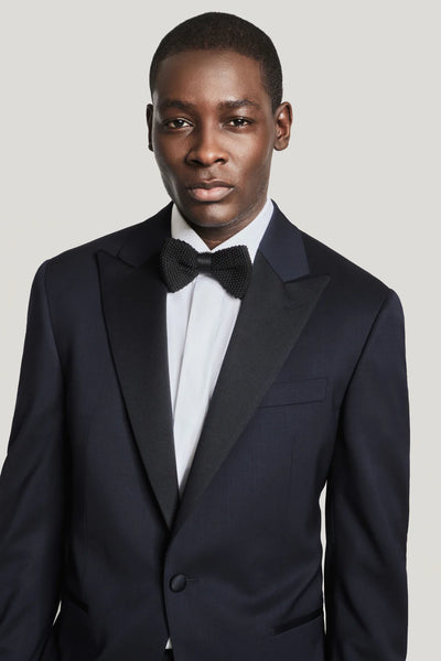 A man poses against a plain light background wearing a Jack Victor Elwood Solid Tuxedo with a peak lapel, white dress shirt, and black bow tie.