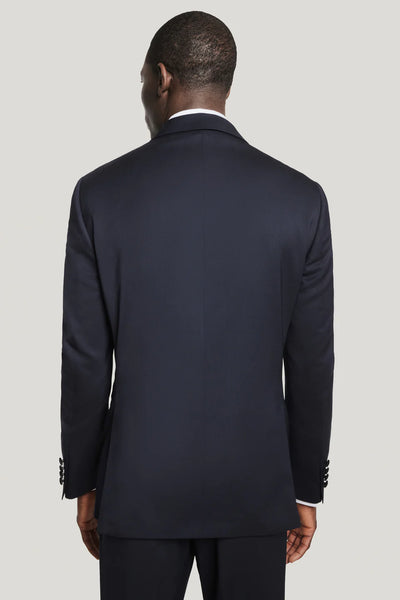 A man is seen from behind wearing a Jack Victor Elwood Solid Tuxedo in dark navy with peak lapel and matching pants, crafted from luxurious Super 130’s wool by Jack Victor, set against a plain light background.