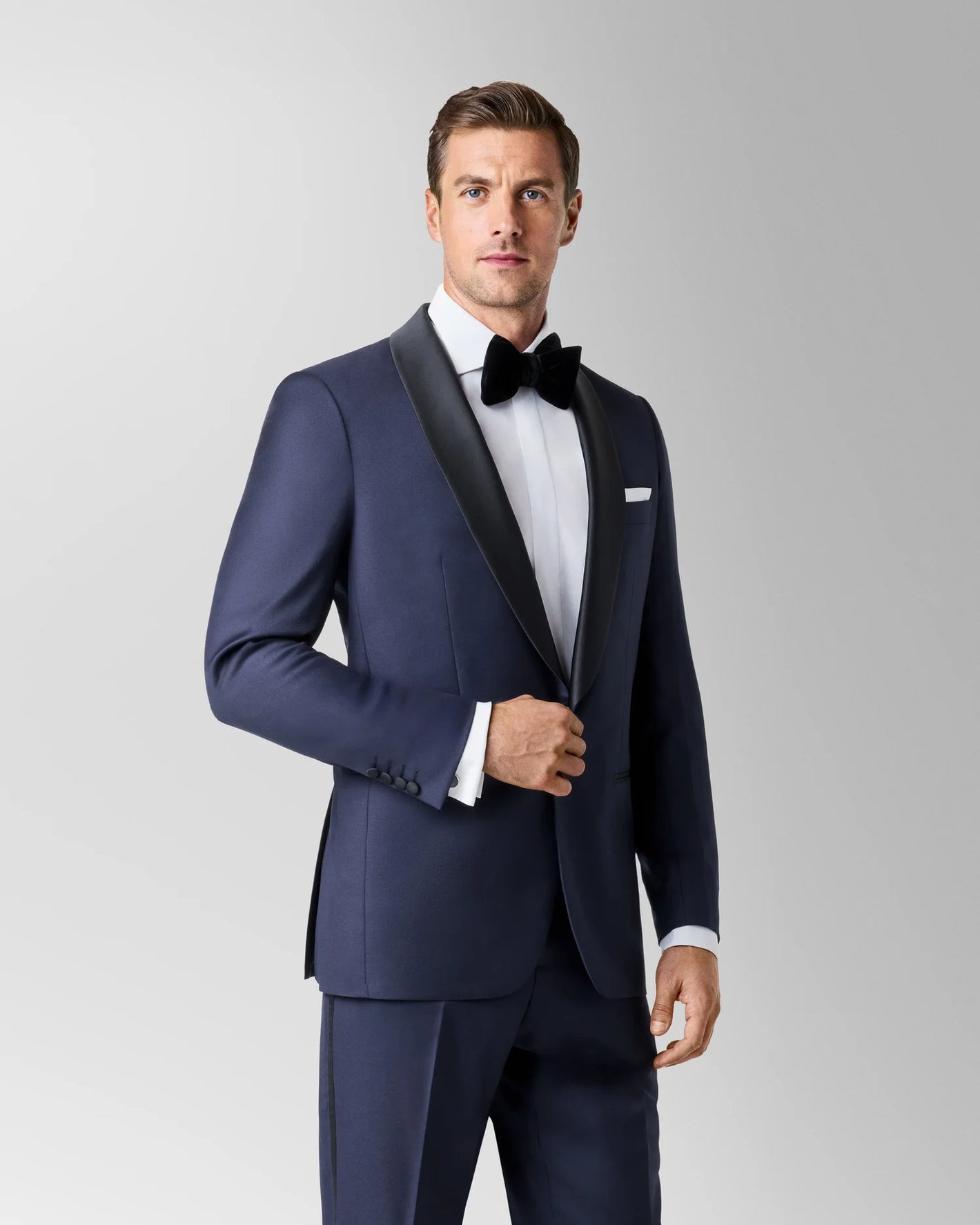A man stands against a plain light background wearing the Baroni MaxDavoli Blue Tuxedo—Italian wool with black satin shawl lapels—paired with a white dress shirt, black bow tie, and pocket square.