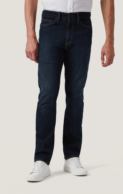 A person wearing 34 Heritage Dark Comfort straight leg jeans from 34 Heritage, paired with a white shirt and white sneakers, stands against a plain light background.