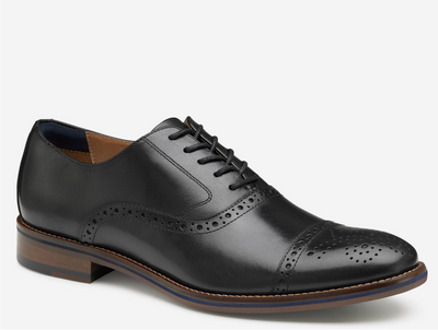 The Johnston & Murphy Conard 2.0 Cap Toe is a black leather men’s Oxford dress shoe with brogue detailing, cushioned comfort, and a brown sole, pictured on a white background.