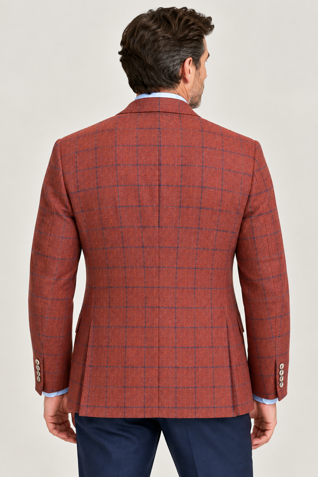 A man is seen from behind wearing the TailoRed Garro Rust Sport Coat 8110694 by TailoRed—a rust-colored, windowpane plaid blazer crafted from fine Italian wool—paired with blue trousers and set against a plain backdrop, making it a perfect men’s fall jacket.