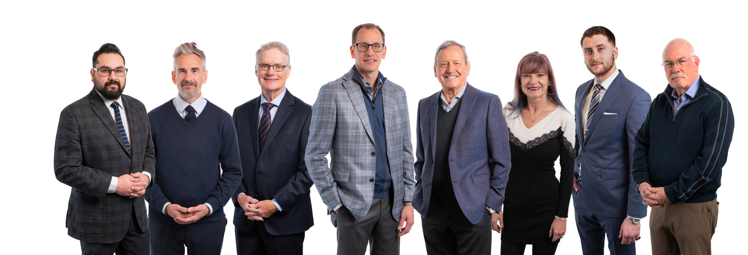 A group of eight adults, six men and two women, stand in a row facing the camera, all dressed in business or smart casual attire against a white background.