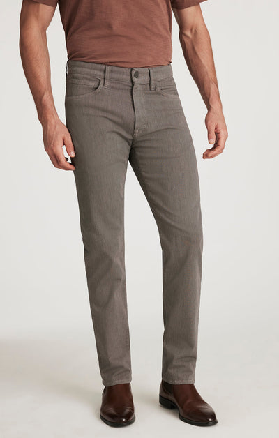 Wearing a brown short-sleeve shirt, 34 Heritage Courage Diagonal Pants by 34 Heritage in a straight fit, and brown dress boots, a person stands against a light plain background.