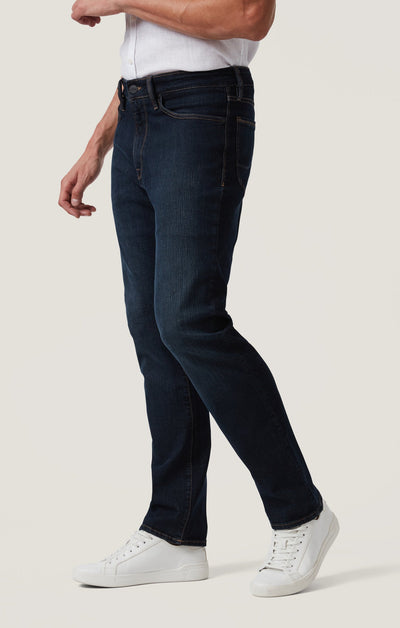 A person wears 34 Heritage Dark Comfort jeans by 34 Heritage—dark blue, high-rise, straight-leg five-pocket pants—paired with a white shirt and sneakers, standing against a plain light background.