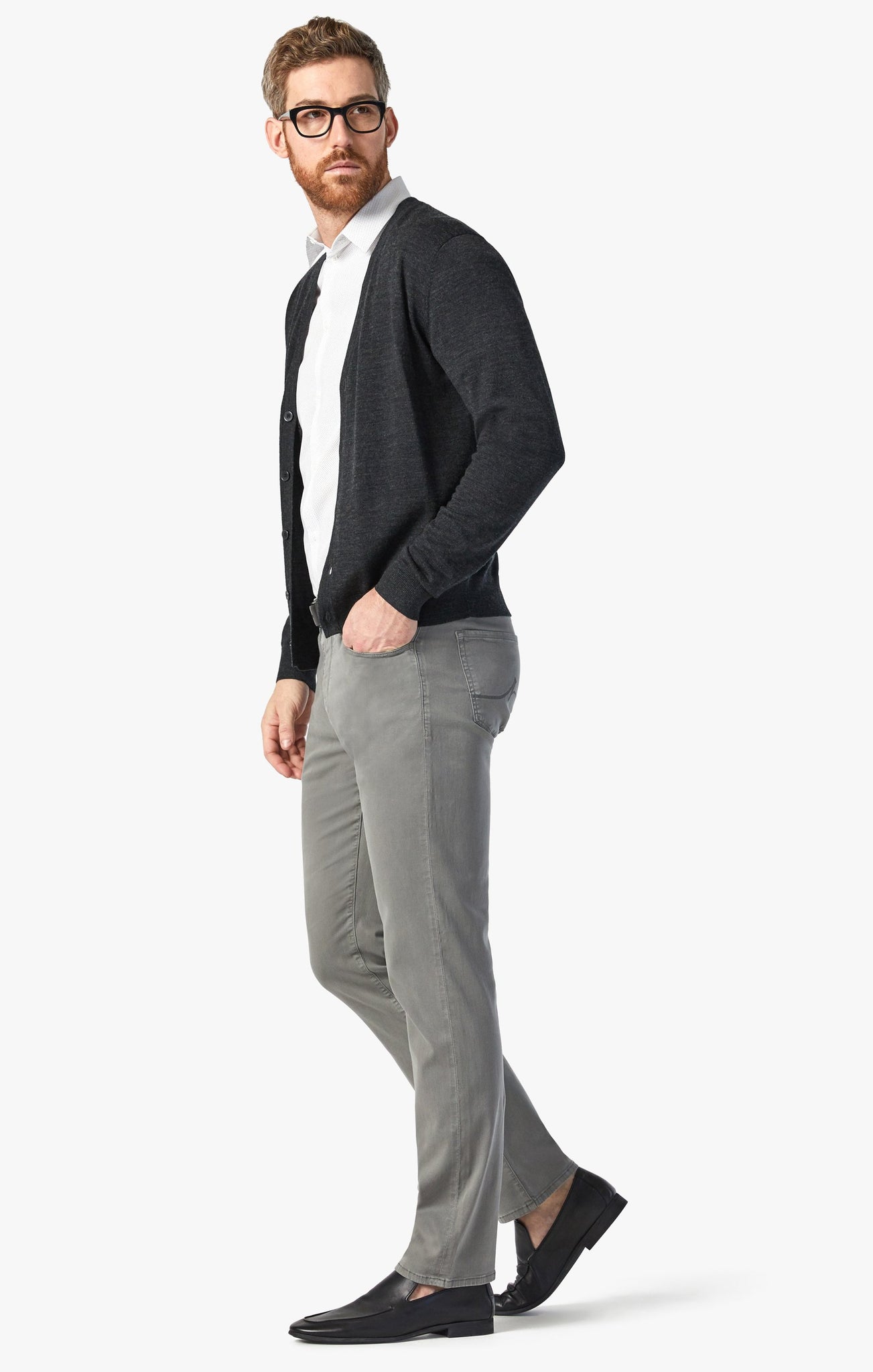 A man in a black cardigan, white shirt, 34 Heritage Pewter Twill relaxed straight-leg gray pants, and black loafers stands against a plain white background.