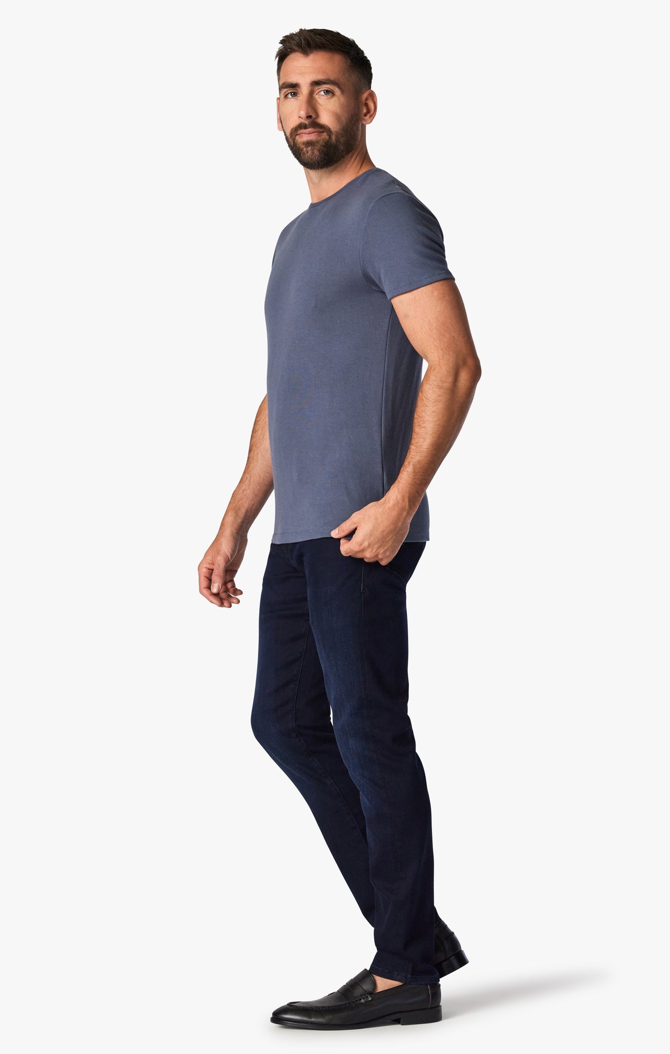 A bearded man in a blue t-shirt, 34 Heritage Ink Urban modern straight-leg dark-wash jeans by 34 Heritage, and black loafers stands facing the camera against a white background.