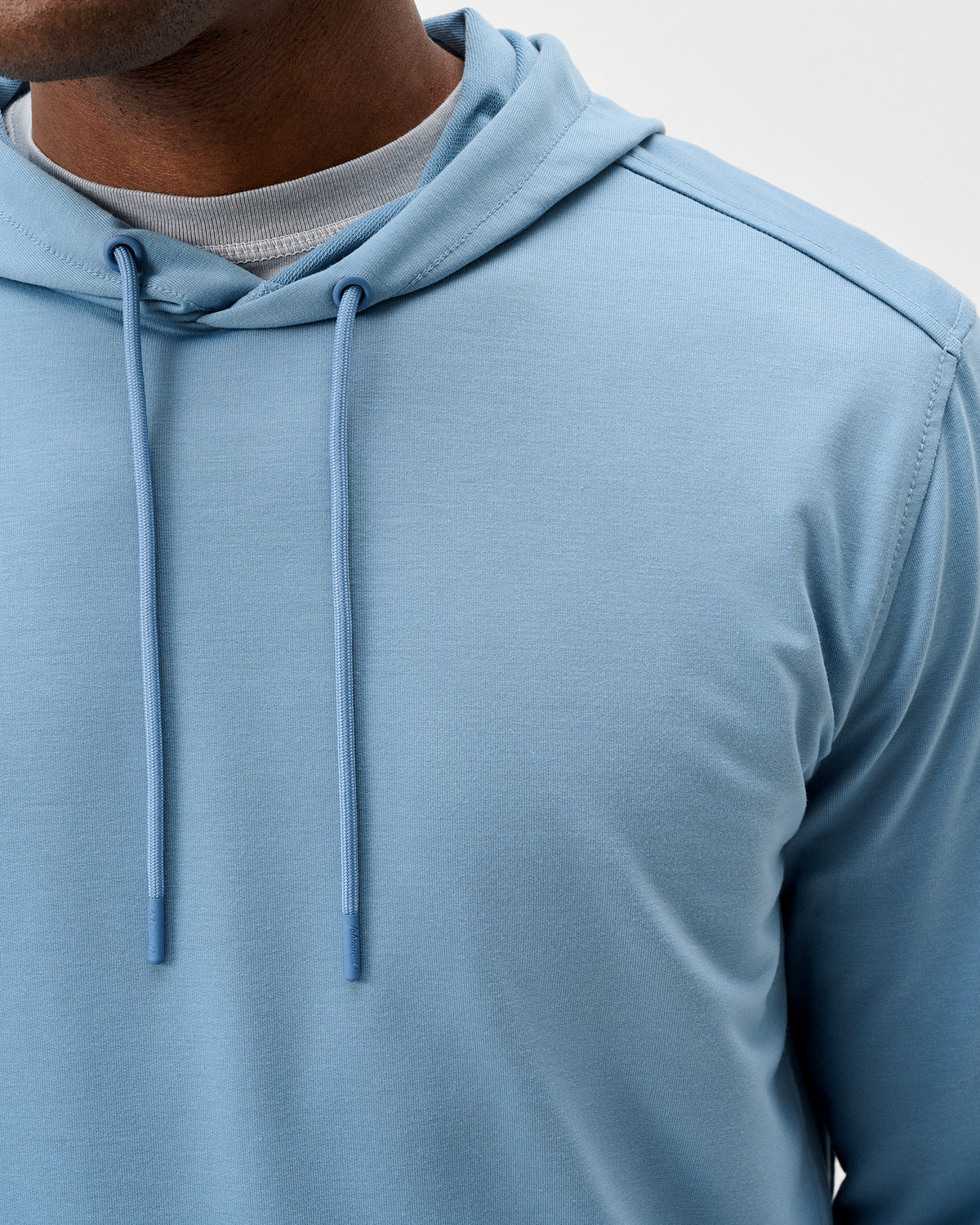 A person wearing the Johnnie-O Willard Performance Hoodie Big & Tall in light blue, featuring drawstrings and layered over a gray shirt, is shown from the chest up. This athletic fit hoodie by Johnnie-O offers moisture-wicking fabric for all-day comfort.