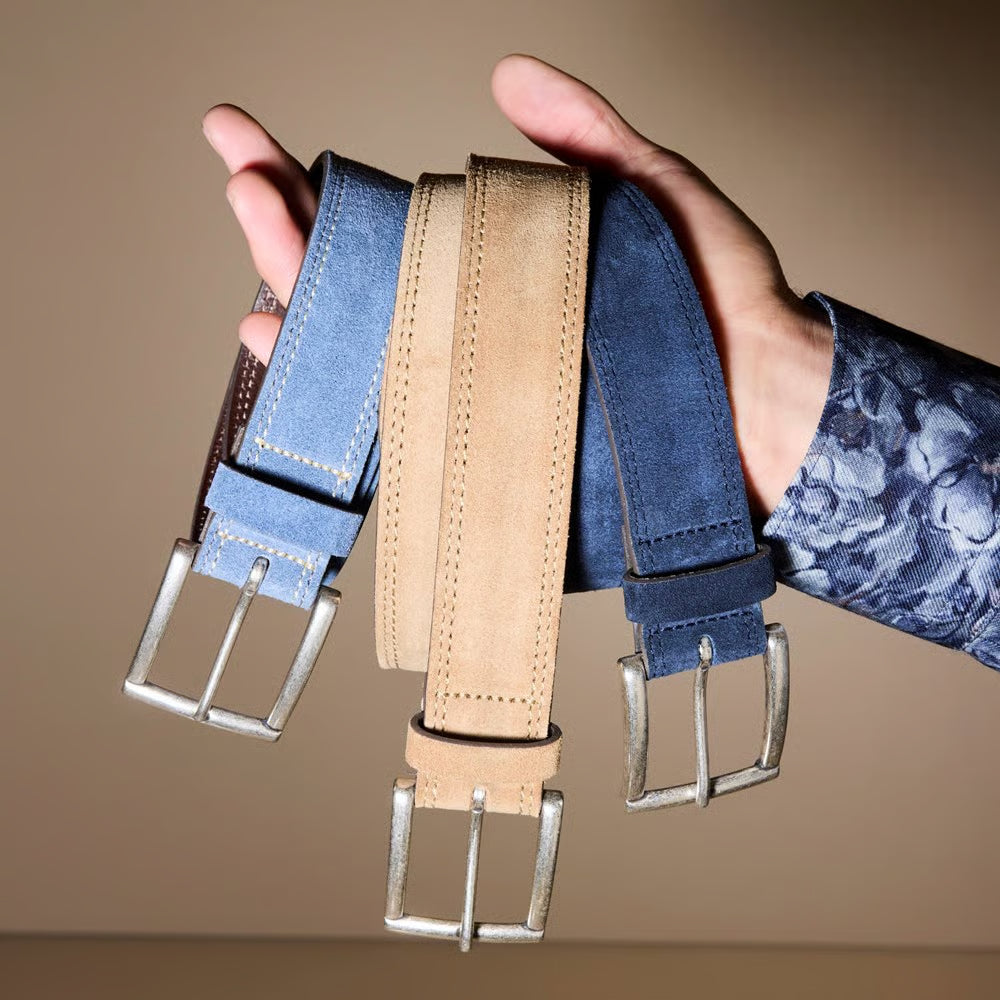 An individual wearing a patterned shirt holds three Allen Edmonds Bourbon Street Suede Belts—each handcrafted in America by Allen Edmonds from premium European suede in blue and beige, featuring elegant silver buckles—all displayed against a plain background.