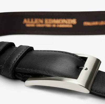 Close-up of the Allen Edmonds Wide Basic Belt in black leather featuring a silver buckle. The inner side displays the Allen Edmonds brand in gold lettering, emphasizing its refined Italian calfskin craftsmanship.