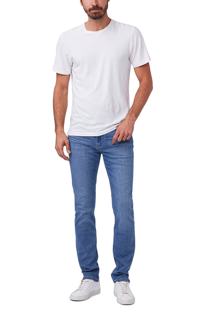 A man stands against a white background wearing a plain white t-shirt, Paige Federal Straight Slim Jeans by Paige in medium wash, and white sneakers.