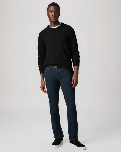 A man stands against a plain background wearing a black sweater, Paige Federal Straight Slim Jeans by Paige in a dark wash, and black slip-on shoes.