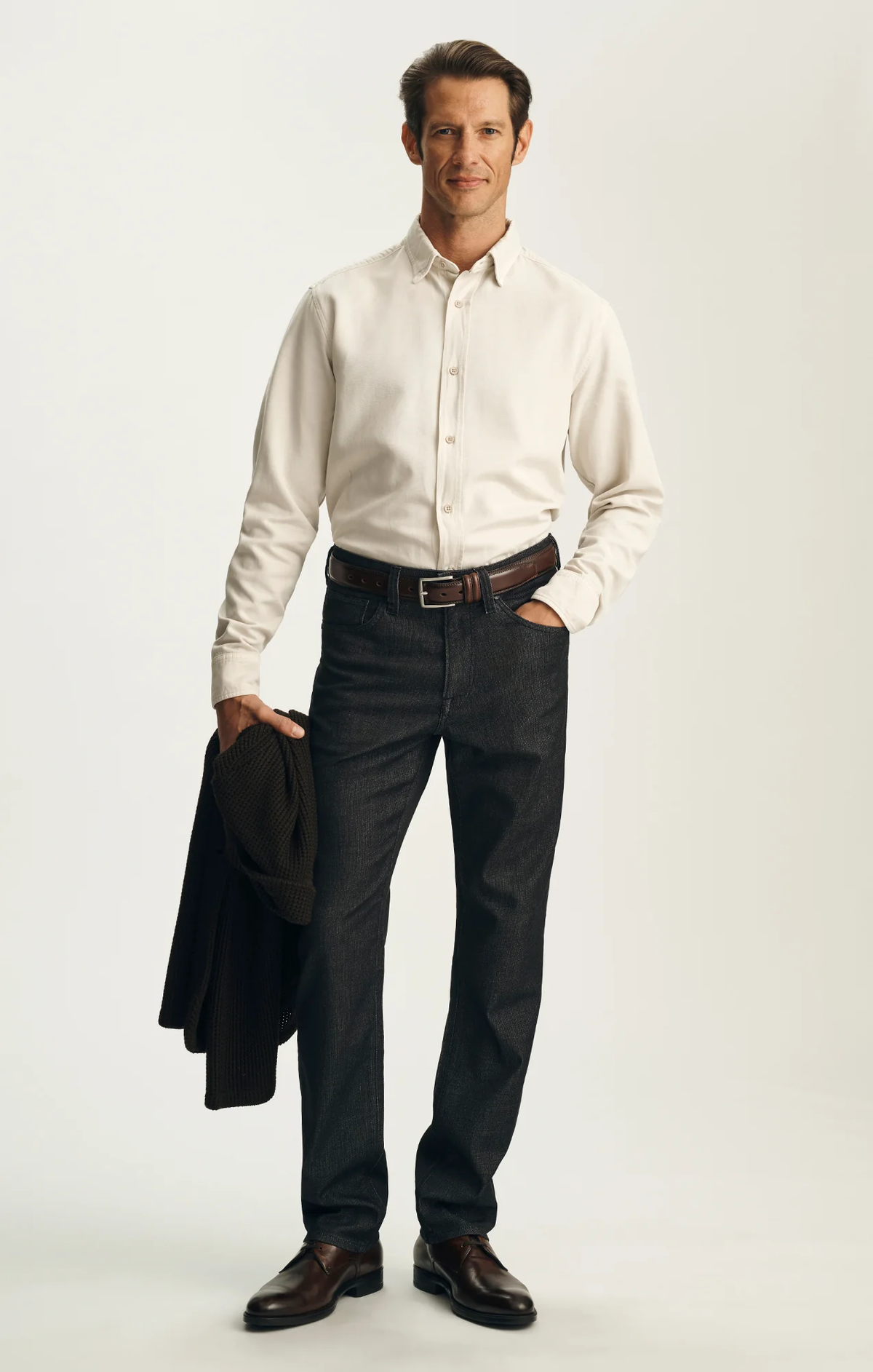 A man stands against a plain background wearing the 34 Heritage Charisma Sporty Big&Tall trousers in a dark wash, paired with a cream button-down shirt, brown shoes, and holding a brown jacket.