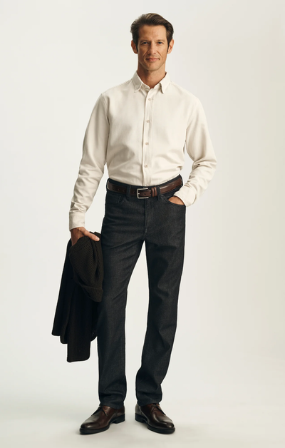 A man stands against a plain background wearing the 34 Heritage Charisma Sporty Big&Tall trousers in a dark wash, paired with a cream button-down shirt, brown shoes, and holding a brown jacket.
