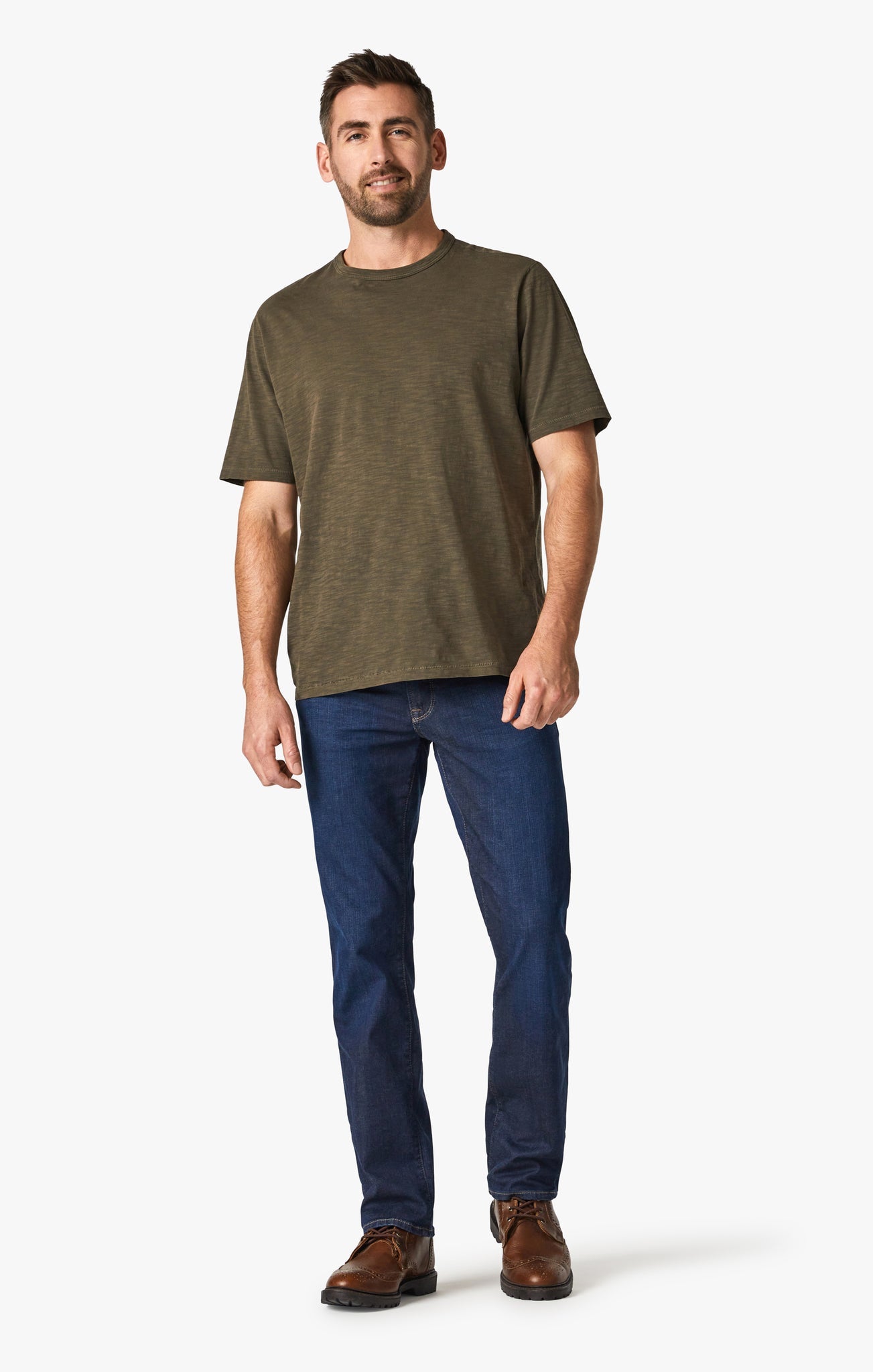 A man stands facing forward against a plain white background, wearing 34 Heritage Dark Cashmere high rise straight leg jeans in soft denim, an olive green short-sleeve t-shirt, and brown shoes.