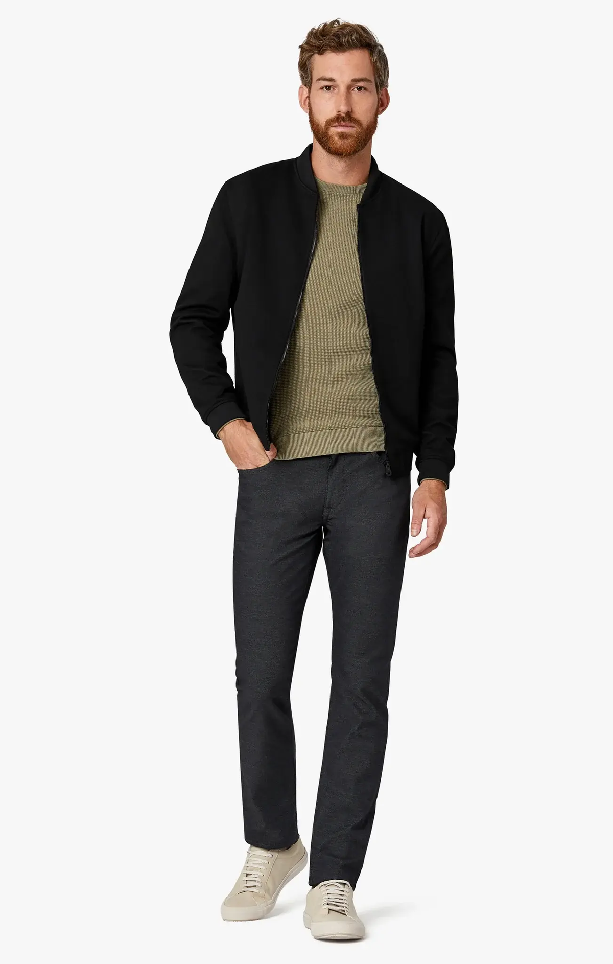 A man wearing a black bomber jacket over an olive green sweater, 34 Heritage Courage Coolmax straight leg trousers, and light-colored sneakers.