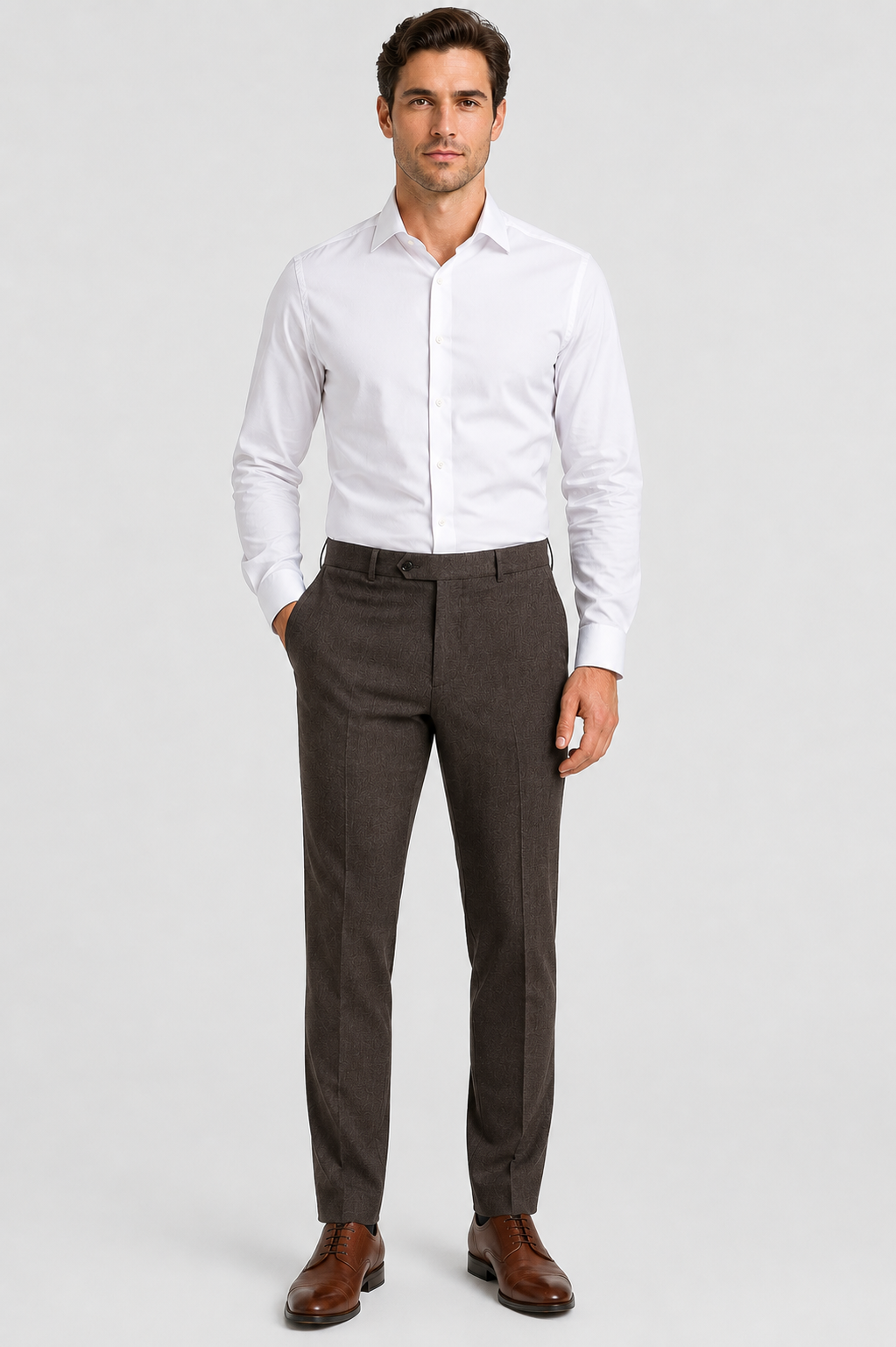 A man stands against a plain background, dressed in a crisp white dress shirt and Ballin M5549 Soho Trousers Seasonal by Ballin—luxurious brown trousers expertly tailored from Super 120’s wool gabardine—paired with matching brown dress shoes. With one hand casually in his pocket, he exudes refined Comfort-“EZE” style.
