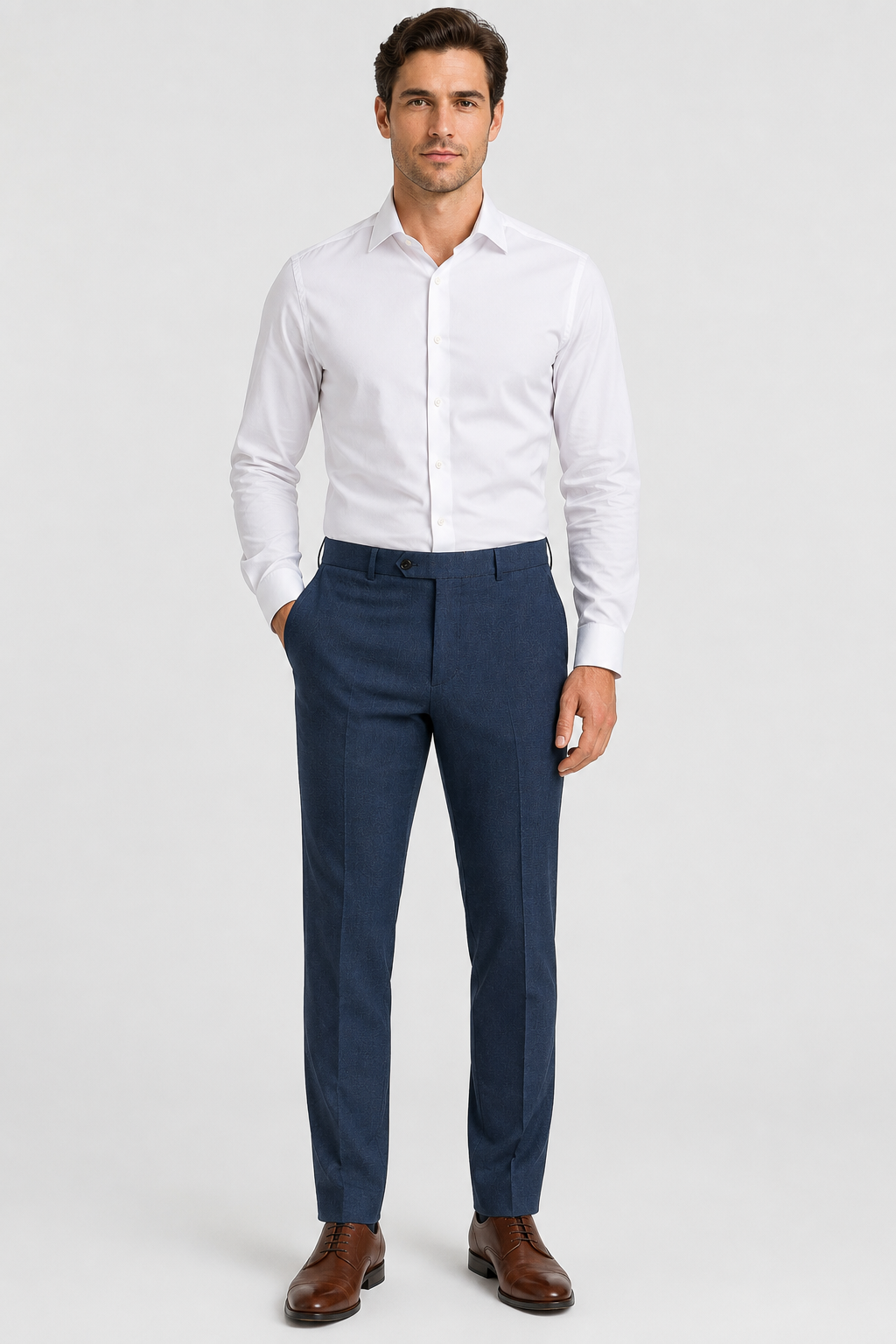 A man stands against a plain background, wearing a white dress shirt, brown dress shoes, and Ballin M5549 Soho Trousers Seasonal by Ballin in blue Super 120’s wool gabardine, with his hands in his pockets.