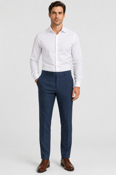 A man stands against a plain background, wearing a white dress shirt, brown dress shoes, and Ballin M5549 Soho Trousers Seasonal by Ballin in blue Super 120’s wool gabardine, with his hands in his pockets.