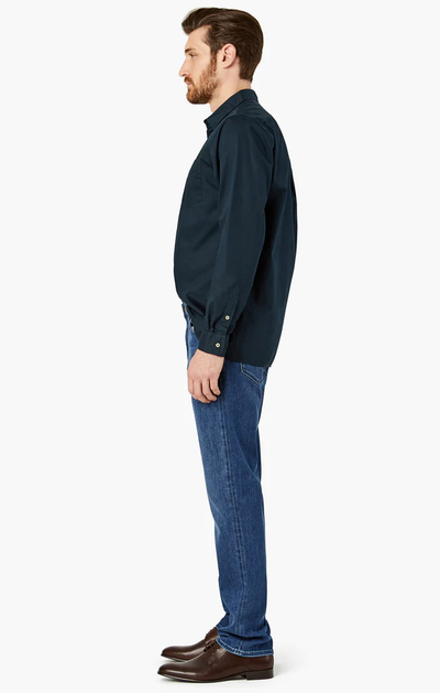 A man in profile wears 34 Heritage Charisma Mid Urban jeans and a navy long-sleeve shirt with dark brown dress shoes, set against a white background.