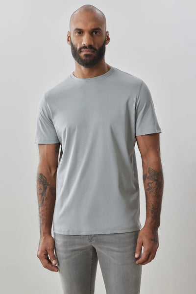A bearded, tattooed man wears a light grey Robert Barakett Georgia Crew 23336 tee and grey jeans, standing against a plain light background.