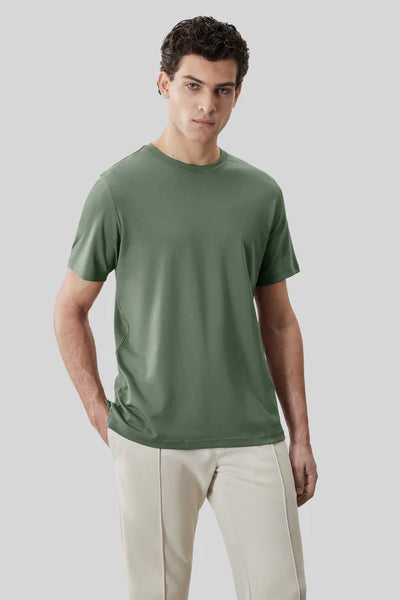 A man stands against a plain grey background wearing a sage green Robert Barakett Tee Shirt Georgia Crew 23336 and light beige pants, with one hand in his pocket.