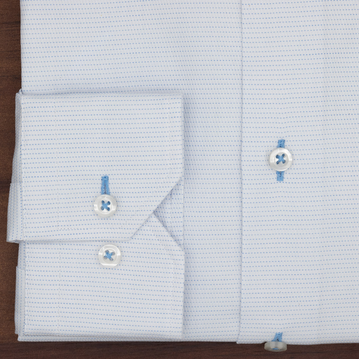 The Hagen Micro Dot Shirt MB7940100 by Hagen is a light blue jacquard dress shirt featuring a subtle micro dot pattern, folded to display its stylish cuff. Two white buttons accented with blue thread and part of the front placket are visible against a wooden surface. Crafted from 100% cotton, this shirt exemplifies modern fit menswear at its best.