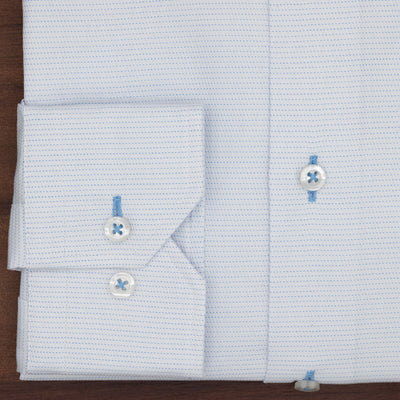 The Hagen Micro Dot Shirt MB7940100 by Hagen is a light blue jacquard dress shirt featuring a subtle micro dot pattern, folded to display its stylish cuff. Two white buttons accented with blue thread and part of the front placket are visible against a wooden surface. Crafted from 100% cotton, this shirt exemplifies modern fit menswear at its best.