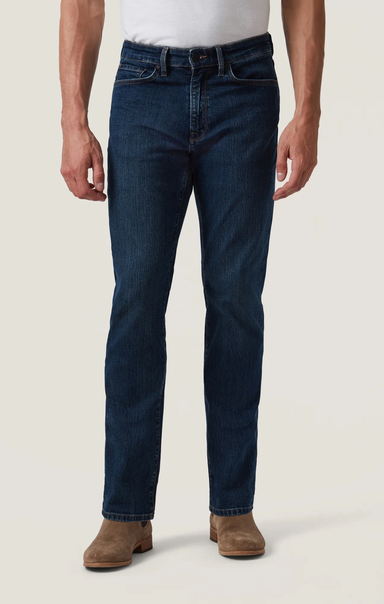 Wearing 34 Heritage Charisma Mid Comfort pants, a white shirt, and light brown suede boots, a person stands against a plain background.