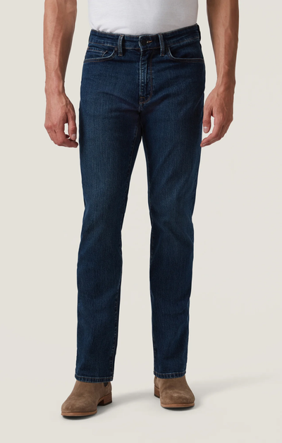 Wearing 34 Heritage Charisma Mid Comfort pants, a white shirt, and light brown suede boots, a person stands against a plain background.