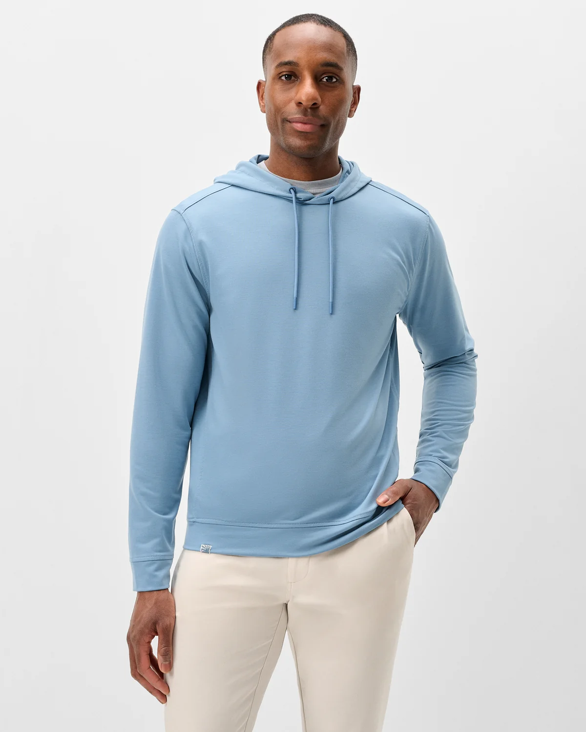 A man stands against a plain background wearing the Johnnie-O Willard Performance Hoodie Big & Tall in light blue, featuring an athletic fit, paired with off-white pants and one hand casually in his pocket.
