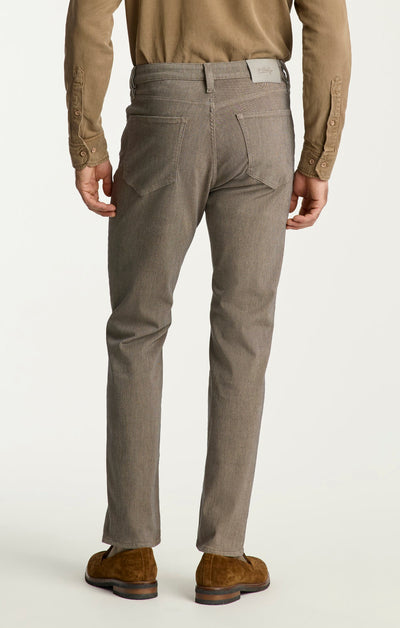 A person wearing 34 Heritage Charisma Diagonal Jeans Big & Tall, a tan shirt, brown belt, and brown suede shoes, shown from the back against a plain background.