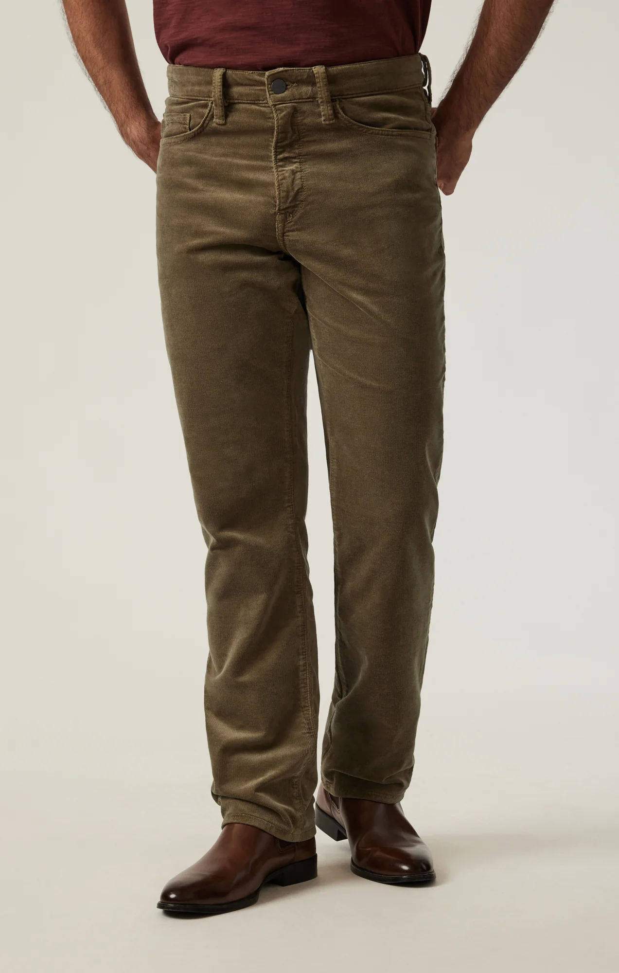 A person wears 34 Heritage Charisma Cord olive green corduroy pants, a maroon shirt, and brown leather boots while standing against a plain light background.