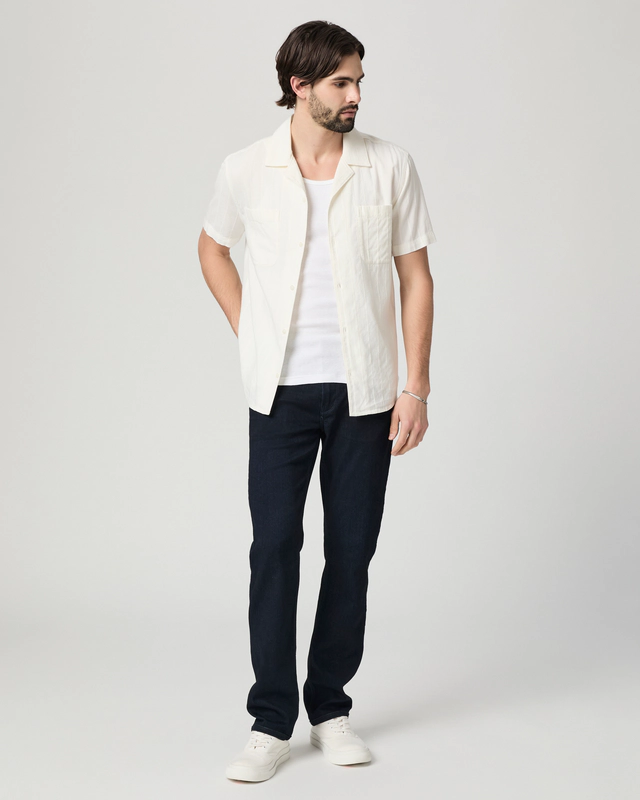 A man wearing Paige Federal Straight Slim Jeans by Paige, a white short-sleeve shirt over a white tee, and white sneakers stands against a plain light background, looking to his right.