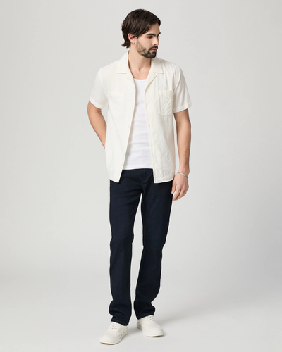 A man wearing Paige Federal Straight Slim Jeans by Paige, a white short-sleeve shirt over a white tee, and white sneakers stands against a plain light background, looking to his right.