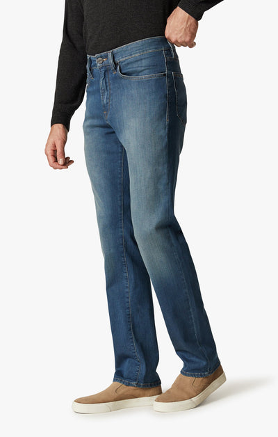 Wearing 34 Heritage Mid Cashmere jeans by 34 Heritage, a person in a black sweater and tan slip-ons stands with one hand in their pocket against a plain white background.