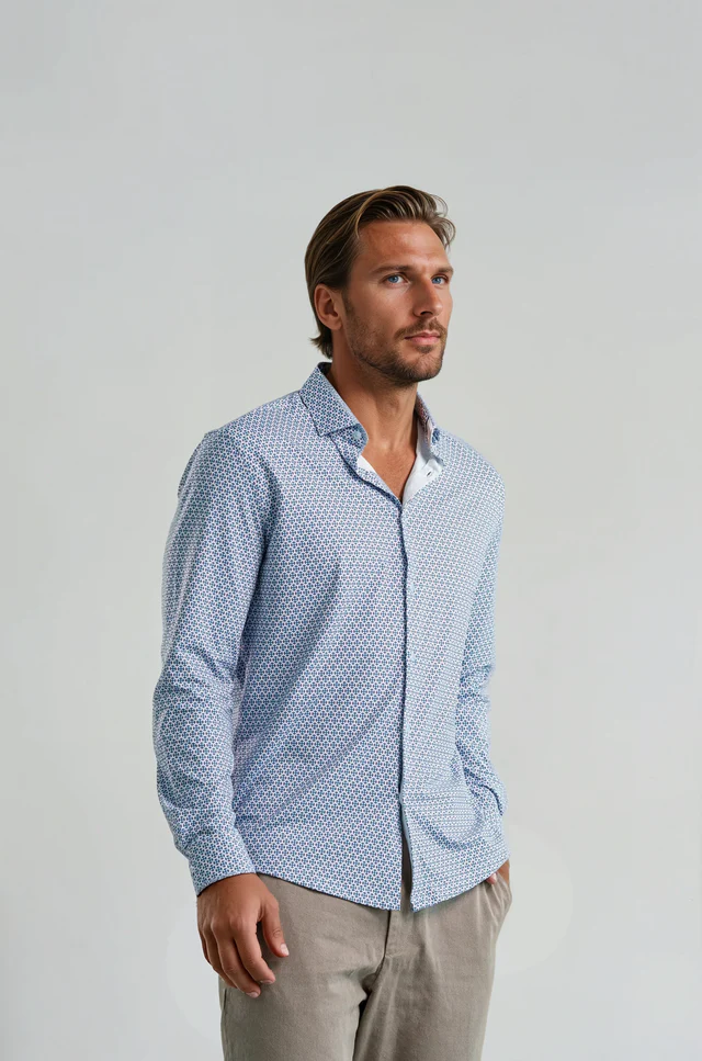 A man stands facing slightly to the side, wearing a Stone Rose APEX Medallion Geo shirt in light blue with tailored grey trousers against a plain light background.