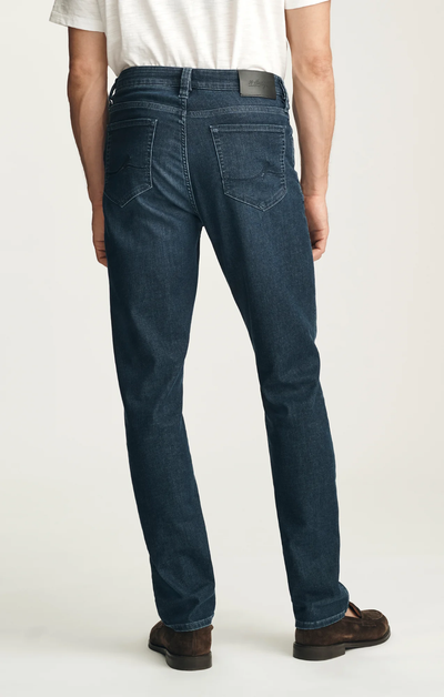 A man stands facing away, wearing a white T-shirt, brown shoes, and 34 Heritage Courage Sporty Jeans in dark blue from 34 Heritage, featuring sporty denim for enhanced comfort, visible back pockets, and a black label on the waistband.