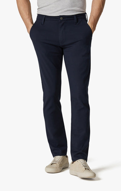 A person wearing a light gray t-shirt, 34 Heritage Night Sky High-Flyer navy slim trousers by 34 Heritage, and beige sneakers stands with hands in pockets against a plain white background.